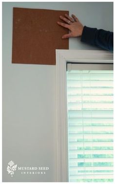 a person holding up a piece of cardboard on top of a window sill next to blinds