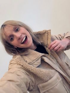 a woman is smiling and holding something in her hand while wearing a tan jacket with sherme