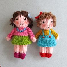 two small knitted dolls are hanging on a white wall, one is wearing a colorful dress and the other has a red flower in her hair