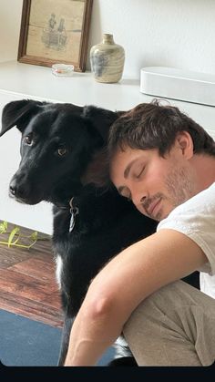 a man sitting on the floor next to a black dog with its head on his chest