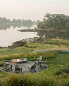 a fire pit surrounded by lawn chairs in the middle of a field with water and trees