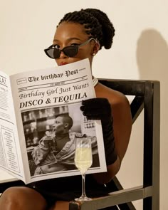 a woman sitting in a chair reading a newspaper with a glass of wine next to her