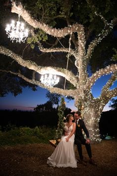 a bride and groom standing under a tree at night with lights on it's branches