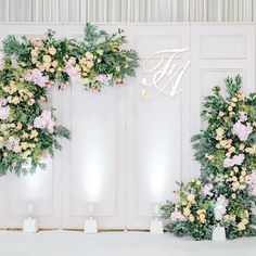 two floral arrangements on display in front of white walls with gold letters and flowers at the top