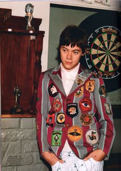 a young man standing in front of a dart board with badges on his jacket and pants