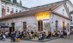 many people are sitting at tables in front of a building with a neon sign on it