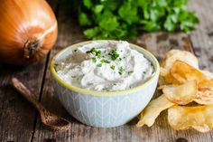 a bowl filled with white dip next to chips and an onion on a wooden table