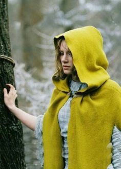 a woman wearing a yellow hooded cape standing next to a tree in the woods,