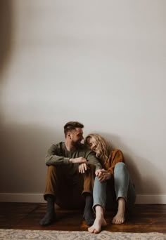 a man and woman sitting on the floor in front of a white wall with their arms around each other