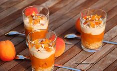 three glasses filled with dessert sitting on top of a wooden table next to two peaches