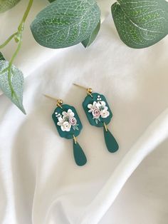 two green earrings with flowers on them sitting next to a white cloth and leafy plant