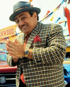 a man wearing a suit and tie standing in front of a car with his hands clasped