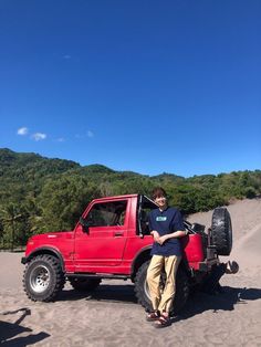 a man standing next to a red truck on top of a sandy hill with trees in the background