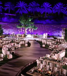 an outdoor event with tables and chairs set up in the grass at night, surrounded by palm trees