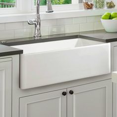 a white kitchen sink sitting under a window next to a bowl of green apples on top of a counter