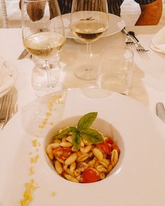 a white plate topped with pasta and veggies next to two glasses of wine