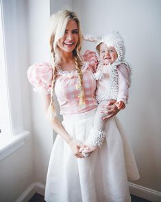 a woman holding a baby in her arms and smiling at the camera while standing next to a window