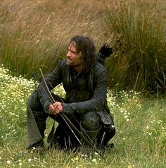 a man with long hair sitting in the grass holding two arrows and looking at something