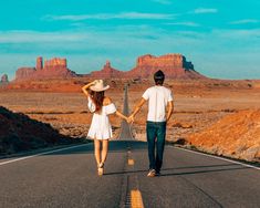 a man and woman holding hands walking down the middle of an empty road with mountains in the background