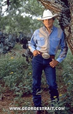 a man wearing a cowboy hat standing next to a tree in the woods with his hands on his hips