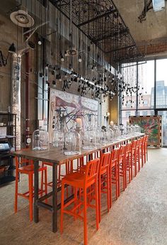 a long table with orange chairs in an industrial style dining room filled with large windows