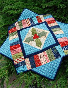 a quilted table topper sitting on the ground in front of some green plants