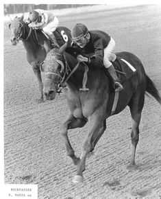 a man riding on the back of a brown horse down a race track in black and white