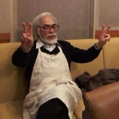 an old man sitting on a couch with his hands in the air and wearing glasses