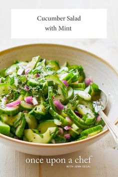 a bowl filled with cucumbers and onions on top of a wooden table next to a spoon