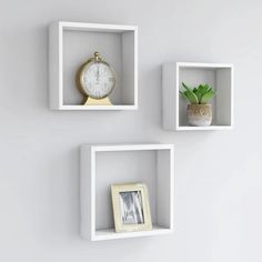 three white square shelves with different types of clocks and plants on them against a gray wall