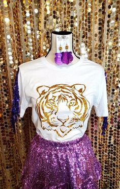 "Game day is for all the girlies! Our one of a kind Tiger Sparkle Tee is designed for cool comfort but with sparkles in mind!  Size inclusive full size run makes sure everyone gets a chance to sparkle! T shirt features: - Bella + Canvas 6400 relaxed fit jersey ring spun cotton feminine tee, but not too form fitting - crew neck - glitter design: gold glitter image, royal purple sequin fringe trim on length of sleeve - coordinating sequin fringe trim Care: wash shirt inside out, preferably in a garment bag on delicate cycle, wash cold. Do not iron design, do not bleach. Line dry or lay flat to dry.  **Any props in photo, including but not limited to accessories, earrings, backdrops, apparel, etc. are for photo purposes only and are NOT included in this listing for purchase.**  Earrings and o Glitter Images, Glitter Design, Royal Purple, Fringe Trim, Game Day, Gold Glitter, Women's Shirt, Womens Clothing Tops, Bella Canvas