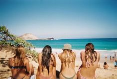 four women in bikinis sitting on the beach looking at the water and people swimming