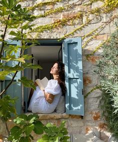 a woman sitting in an open window with a book and looking up at the sky