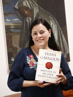 a woman is holding up a book in front of a painting and smiling at the camera