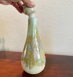 a hand is holding the top of a vase on a wooden table with a white wall in the background