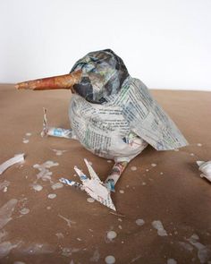 a bird made out of newspaper sitting on top of a brown table covered in paint