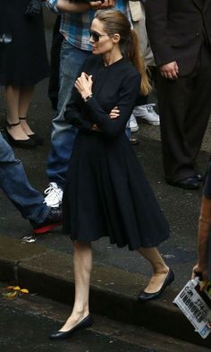 a woman in a black dress and heels standing on the street with her arms crossed