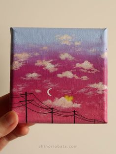 a hand holding up a small painting with the sky and clouds painted in pink, blue, yellow and white