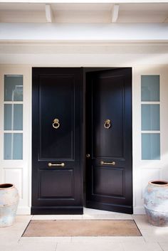 two large black doors are open in front of a white house with vases on the ground