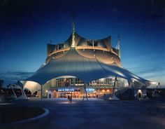 a large blue tent sitting in front of a tall building at night with people walking around it