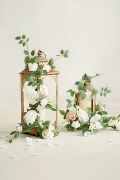 two small lanterns with flowers and greenery on the sides, one is gold and the other is white