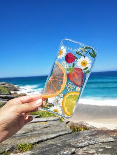 a person holding up a phone case with fruit and flowers on the back in front of an ocean