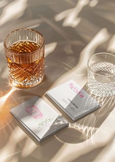 two business cards sitting next to a glass of whiskey on a table with sunlight coming through the window