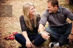 a man and woman sitting on the ground with apples in the background, smiling at each other