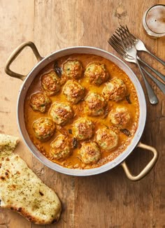 a pan filled with meatballs covered in sauce next to bread and silverware on a wooden table