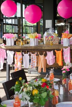 there are many baby clothes hanging on the line outside at this party table with pink and orange balloons in the background