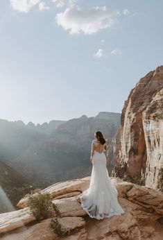 a woman in a wedding dress standing on top of a cliff looking at the mountains