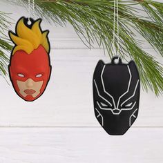 two ornaments hanging from a pine tree on a white wooden background, one has a mask and the other has a face