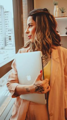 a woman holding an apple laptop in her right hand and looking out the window at the city