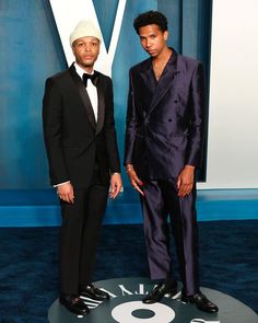 two men in suits standing next to each other on a blue carpet with the letter v behind them